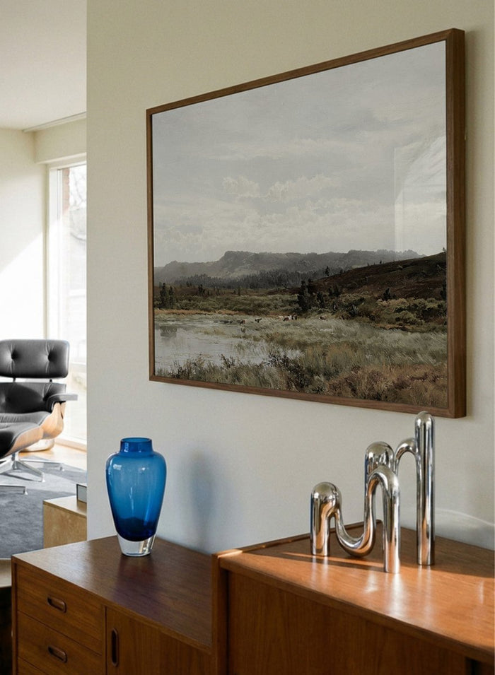 A framed landscape art print depicting a muted, overcast valley scene with a small body of water, grassy foreground, distant hills, and a cloudy sky, shown against a light neutral background. A View of Fleet Moor, Surrey by Alfred de Bréanski is a vintage English landscape print featuring cattle, water, rolling hills, open countryside, and a cloudy sky.