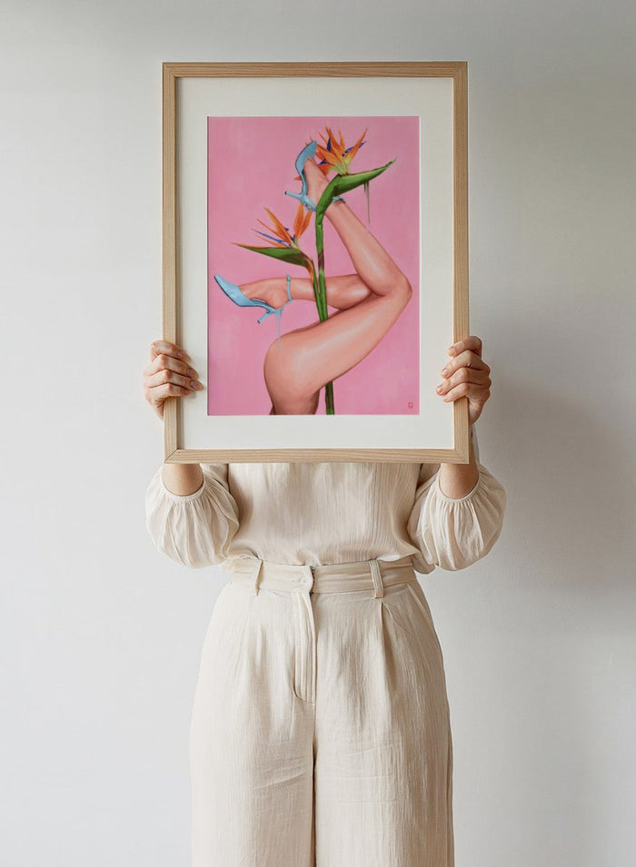 Legs wearing blue high heels with a pink background and bird of paradise flower. Bird of Paradise by Alexander Grahovsky art print featuring crossed legs in blue heels and a bold tropical flower on a pink background. Officially licensed surreal wall poster.