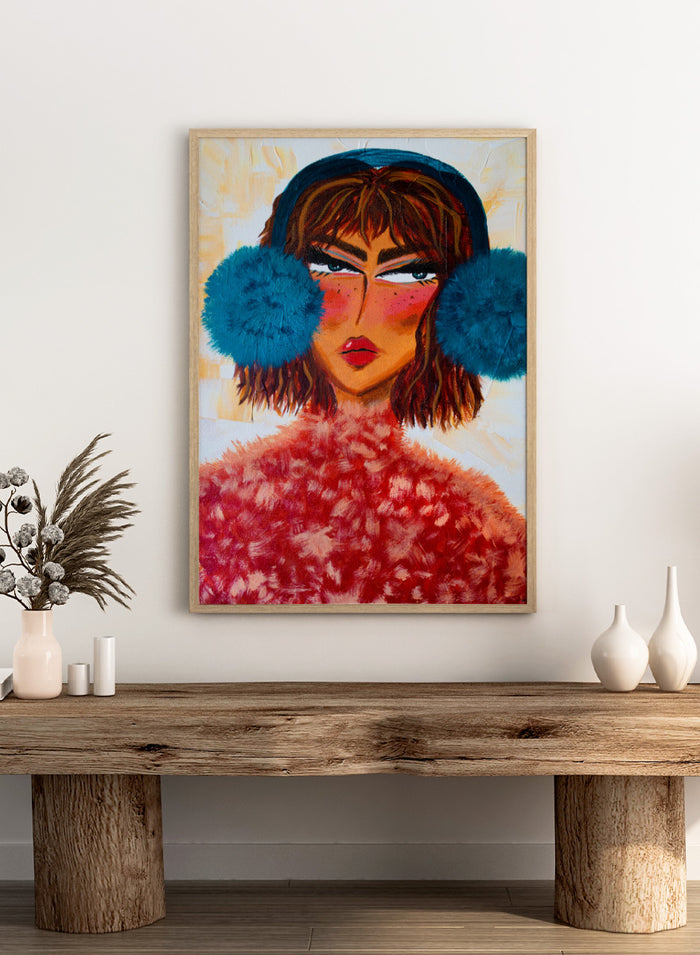 A stylized portrait painting of a woman with brown hair, a dark blue head covering, large bright blue pom-pom ear muffs, and a textured red fuzzy garment, set against a warm cream background. Shop Sassy Earmuffs Portrait by Amy Straley, a contemporary figurative wall art print featuring a bold woman portrait, blue earmuffs, and vivid pink and red tones.