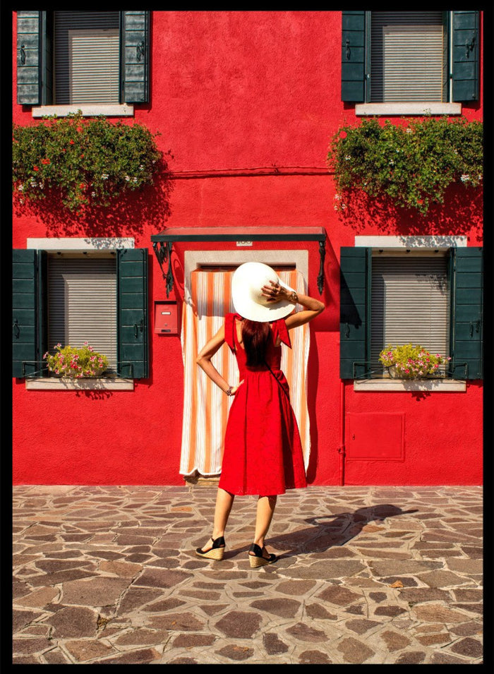 Woman in a red dress and white hat standing in front of a red wall with green shutters. Red Art Print by Anette Ohlendorf featuring a woman in a red dress standing against a red building. Officially licensed travel photography with strong color and symmetry.