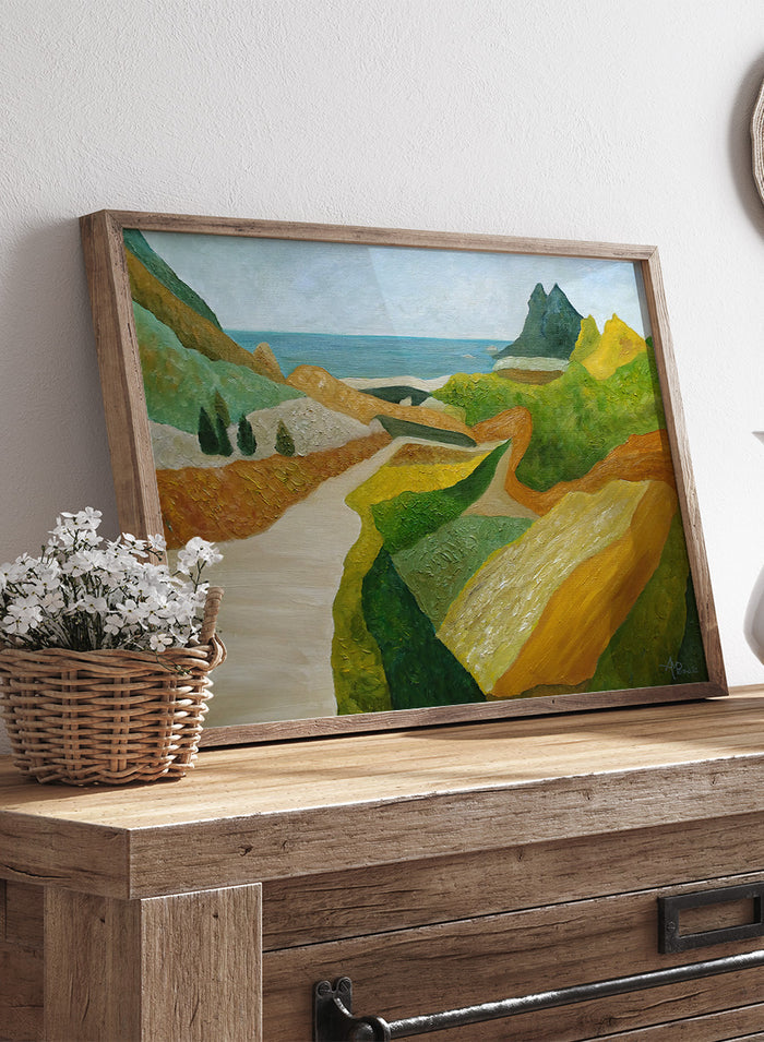 A framed landscape painting featuring a winding cream-colored path through green and ochre hills, leading to a blue sea and distant mountain under a pale sky. Shop Vibrant Coastal Vista by Angeles M. Pomata, a colorful coastal landscape art print featuring winding roads, rolling fields, rugged hills, and blue sea views.