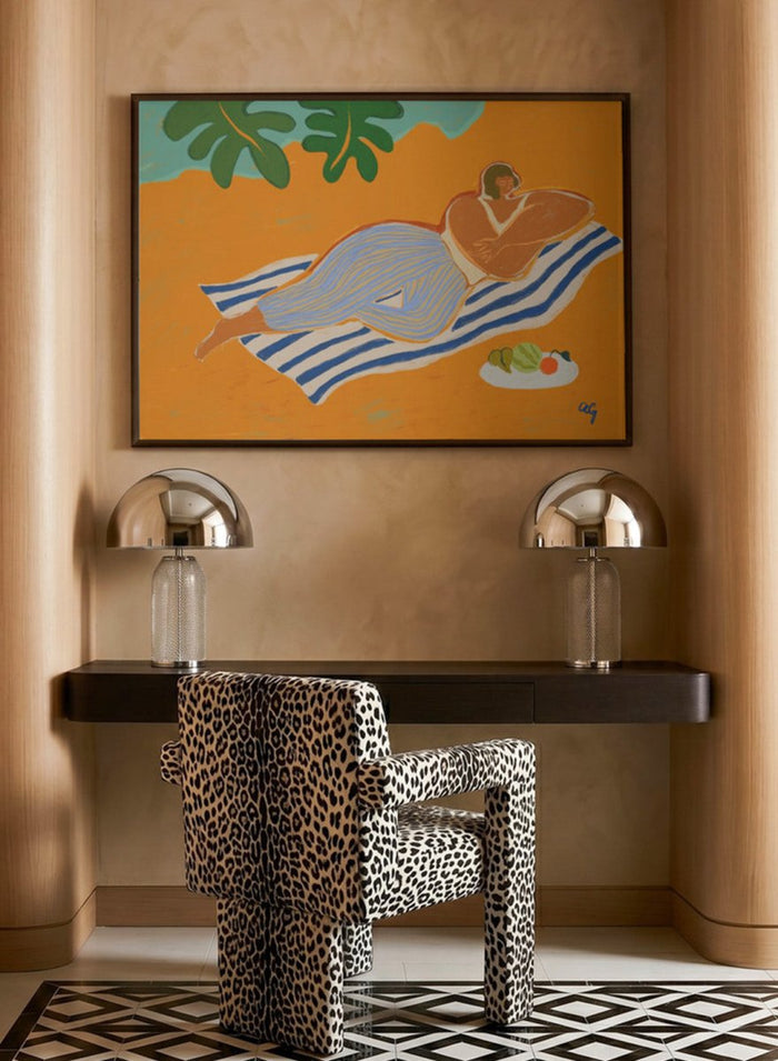 Framed artwork of a woman lounging on a towel with a plate of fruit against an orange background. A Day at the Beach by Arty Guava is a colorful coastal art print featuring a relaxed beach scene, tropical foliage, bold color, and contemporary illustration.