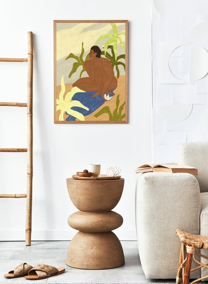 An art print featuring a stylized illustration of a seated figure with dark hair, surrounded by green tropical leaves, a blue cloth, and a white flower, set against a sandy beige and cream background. Au Naturel by Arty Guava premium framed fine art print featuring a seated woman surrounded by oversized tropical plants in warm earthy tones.