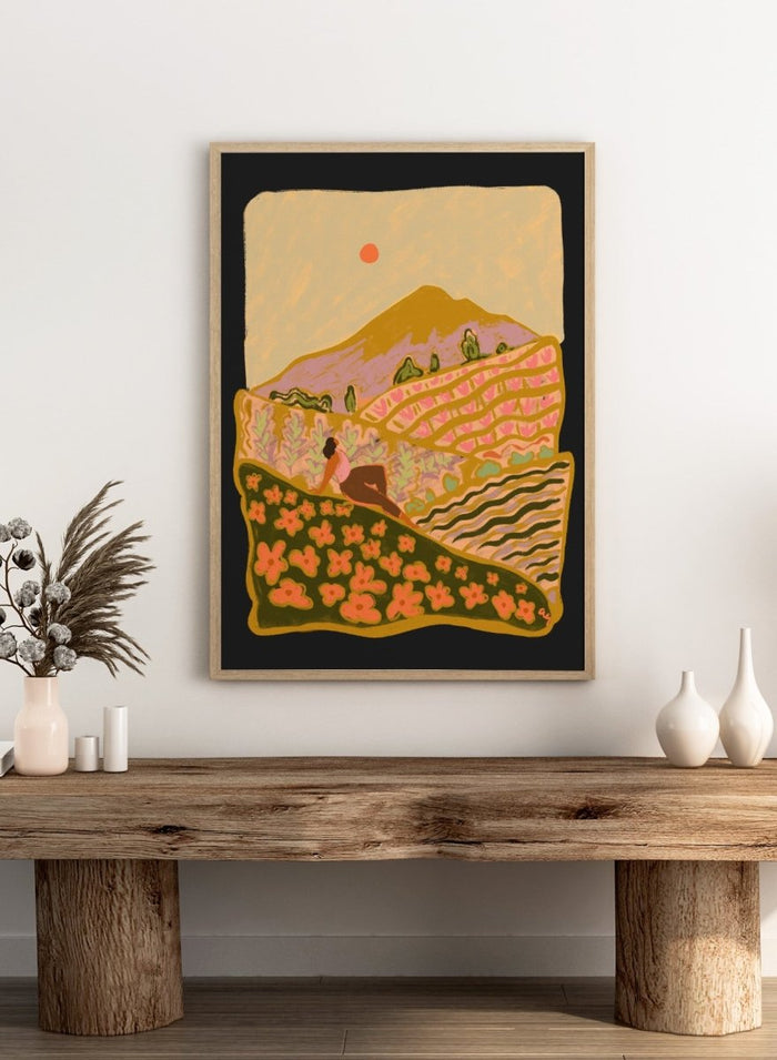 Illustration of a person sitting on a hill with a mountain and sunset in the background. Field Of Flowers by Arty Guava is a colorful landscape art print featuring blooming fields, rolling hills, nature-inspired illustration, and contemporary wall art.