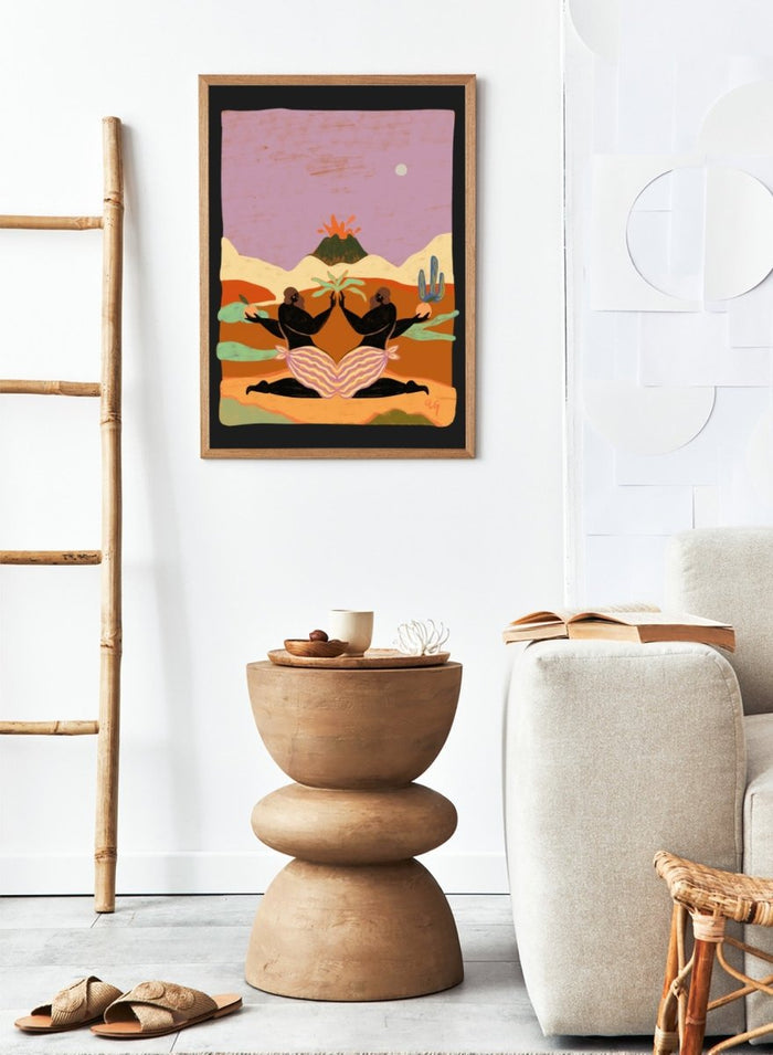 A framed art print featuring a stylized desert landscape with two black silhouetted figures seated in a lotus position, facing each other beneath a volcano and cacti, set against a pink sky. Lava by Arty Guava premium framed fine art print featuring mirrored women, tropical plants, and a glowing volcano in bold, surreal colors.