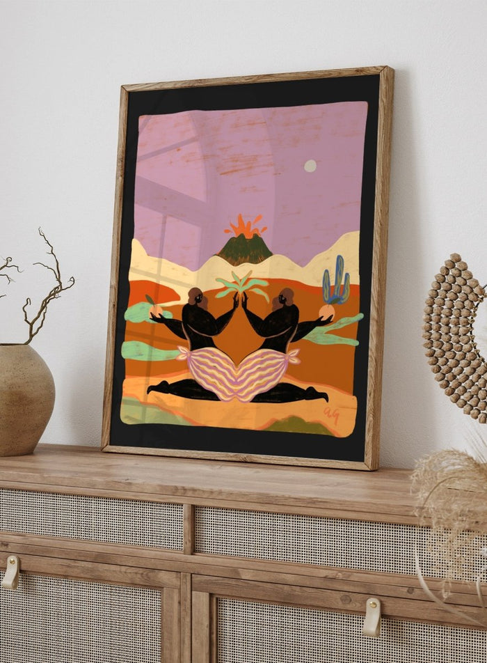 A framed art print featuring a stylized desert landscape with two black silhouetted figures seated in a lotus position, facing each other beneath a volcano and cacti, set against a pink sky. Lava by Arty Guava premium framed fine art print featuring mirrored women, tropical plants, and a glowing volcano in bold, surreal colors.