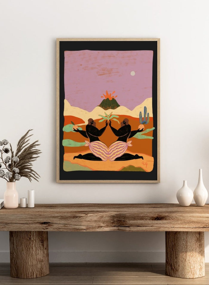 A framed art print featuring a stylized desert landscape with two black silhouetted figures seated in a lotus position, facing each other beneath a volcano and cacti, set against a pink sky. Lava by Arty Guava premium framed fine art print featuring mirrored women, tropical plants, and a glowing volcano in bold, surreal colors.