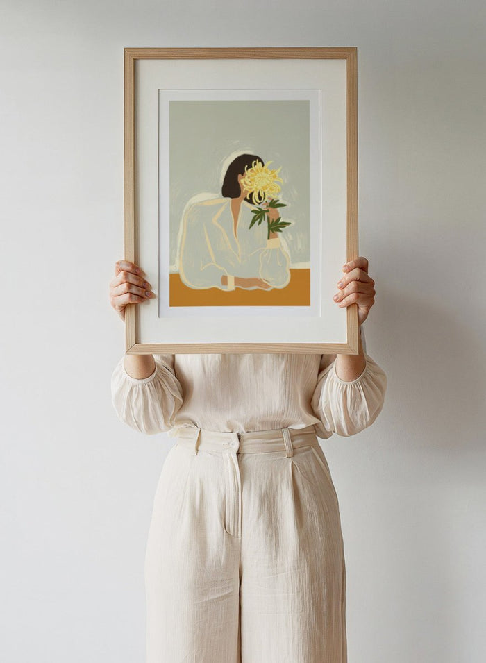 Illustration of a woman in a white dress holding a large yellow flower against a light background. The Chrysanthemum by Arty Guava is a modern portrait illustration featuring a woman with flowers, contemporary floral wall art style, and officially licensed artwork.