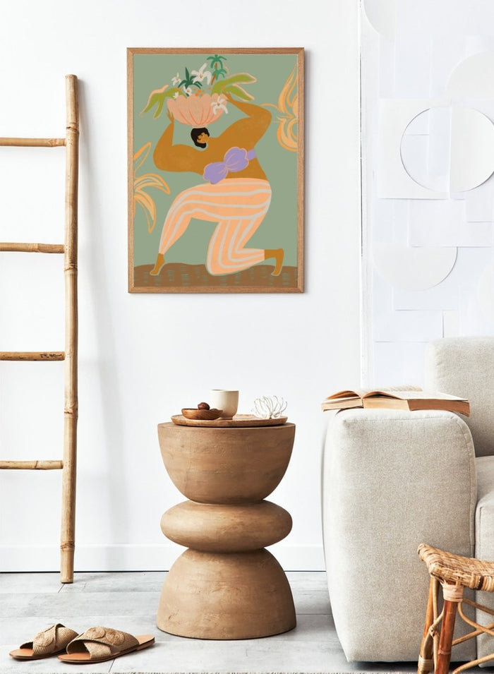 A stylized art print featuring a woman in flowing peach and white striped pants with a purple sash, carrying a large flower basket on her head against a muted sage green background. The Weight of the World by Arty Guava premium framed fine art print featuring a woman carrying tropical flowers in soft green, peach, and lilac tones.