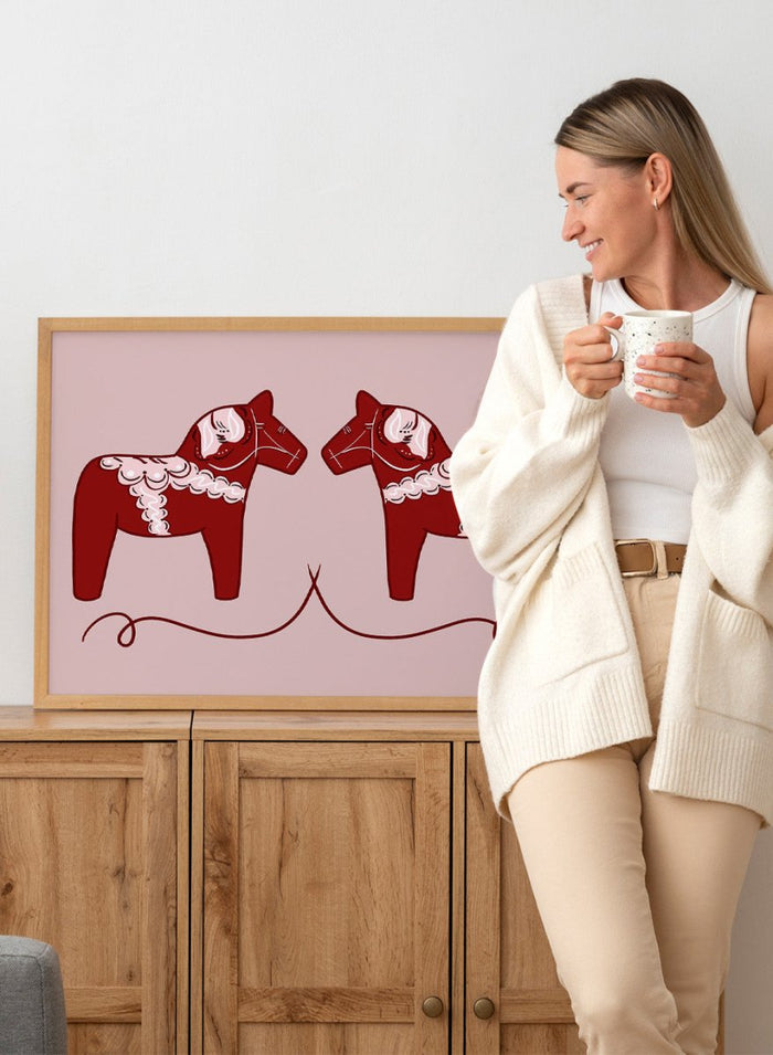 Two red Dala horse figures with white patterns on a pink background. Nordic Horses Twins by Athene Fritsch is a Scandinavian folk art illustration art print featuring Swedish Dala horses, bold red color, and modern wall art design.