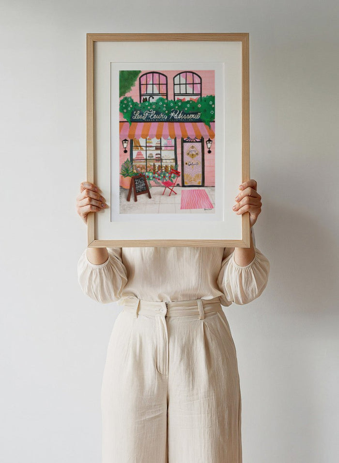 Illustration of a bakery named 'Les Fleuris Patisserie' with a pink exterior and greenery. Bake Shop Front by Baroo Bloom art print featuring a colorful bakery storefront illustration. Whimsical European street scene wall poster with pink tones and playful detail.
