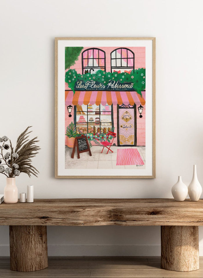 Illustration of a bakery named 'Les Fleuris Patisserie' with a pink exterior and greenery. Bake Shop Front by Baroo Bloom art print featuring a colorful bakery storefront illustration. Whimsical European street scene wall poster with pink tones and playful detail.