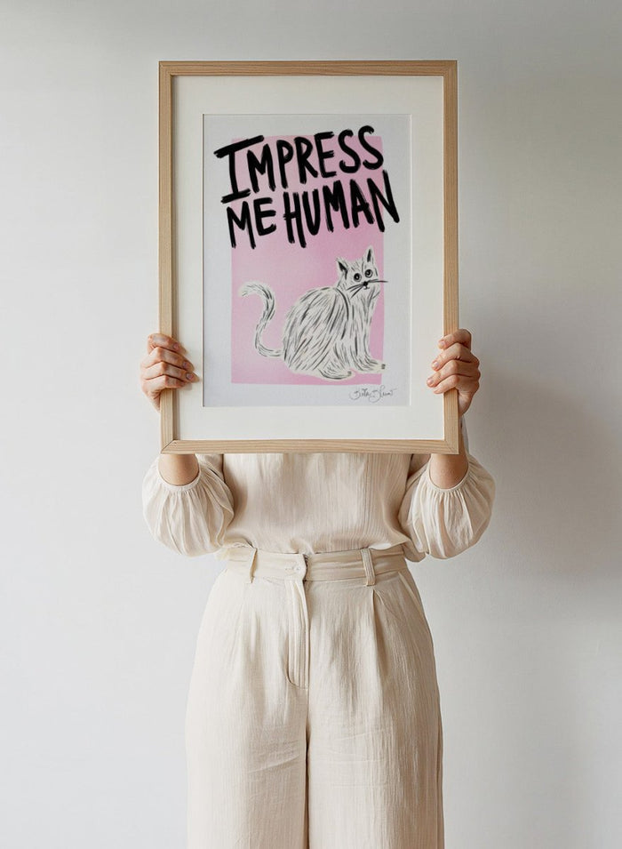 Cartoon cat with 'Impress Me Human' text on a pink background. Cat Owner. Impress Me Human by Baroo Bloom is a playful cat art print featuring hand-drawn illustration and humorous typography. Officially licensed wall poster with optional framing.