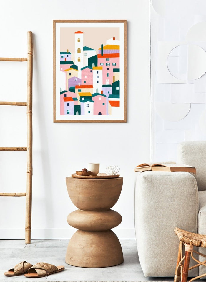 A framed minimalist art print depicting a cluster of stylized pastel houses and a church tower in pink, peach, teal, yellow, and cream tones. Charming Colorful Houses by Bea Müller premium framed fine art print featuring a playful town of pink, lavender, cream, and teal buildings.