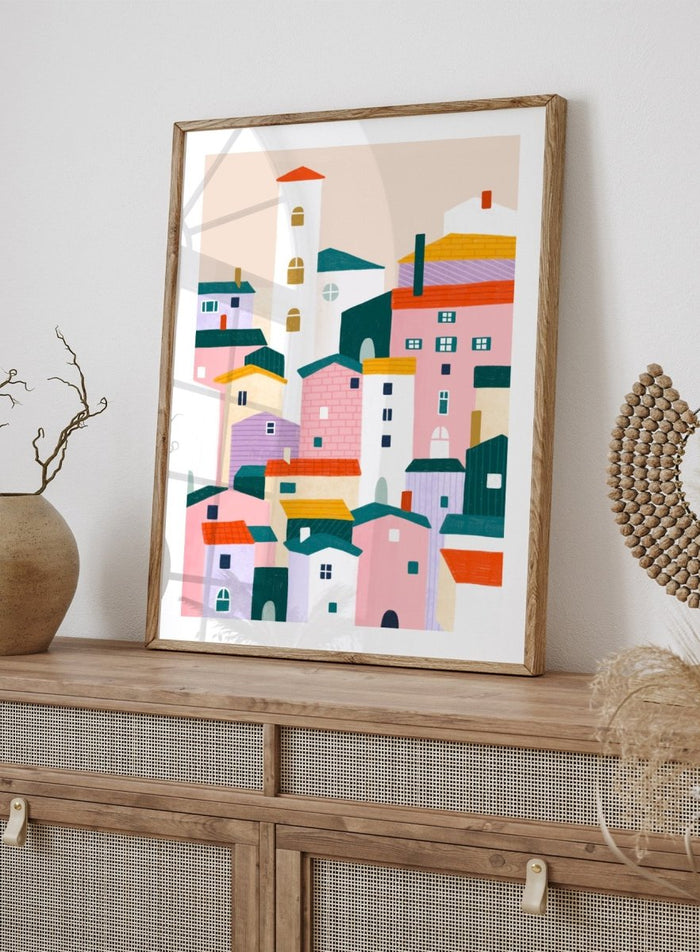 A framed minimalist art print depicting a cluster of stylized pastel houses and a church tower in pink, peach, teal, yellow, and cream tones. Charming Colorful Houses by Bea Müller premium framed fine art print featuring a playful town of pink, lavender, cream, and teal buildings.