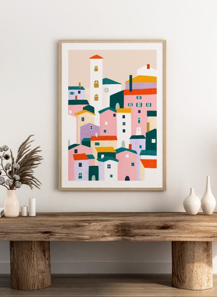 A framed minimalist art print depicting a cluster of stylized pastel houses and a church tower in pink, peach, teal, yellow, and cream tones. Charming Colorful Houses by Bea Müller premium framed fine art print featuring a playful town of pink, lavender, cream, and teal buildings.