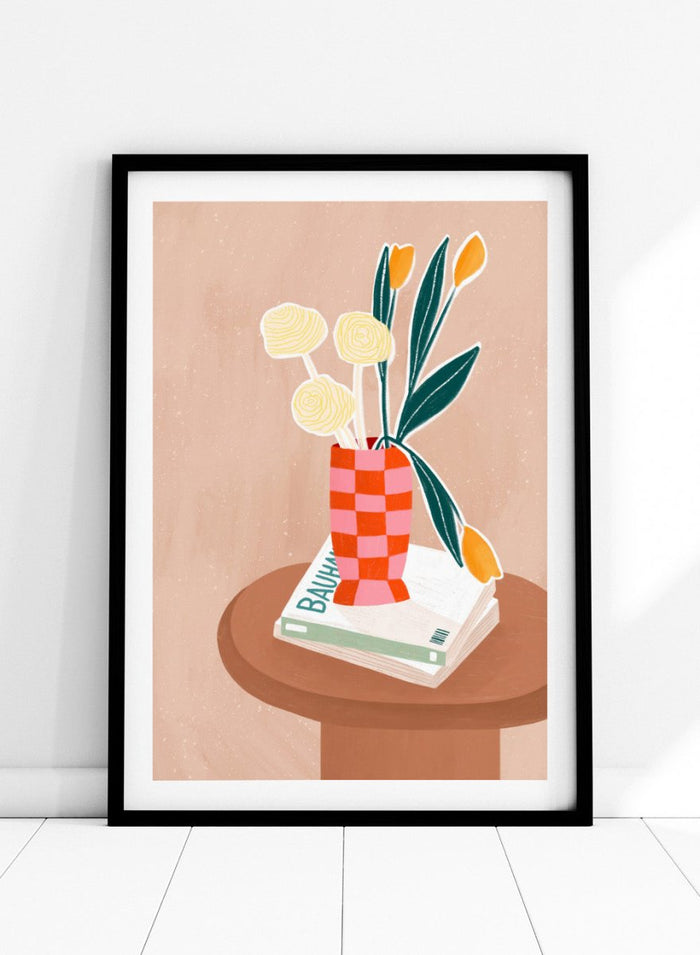 A framed minimalist art print featuring a stylized illustration of a pink and orange checkered vase filled with yellow and orange tulips, resting on a brown round table next to a white book. Colorful Tabletop Flowers by Bea Müller premium framed fine art print featuring yellow tulips and cream blooms in a red-and-pink checkered vase.
