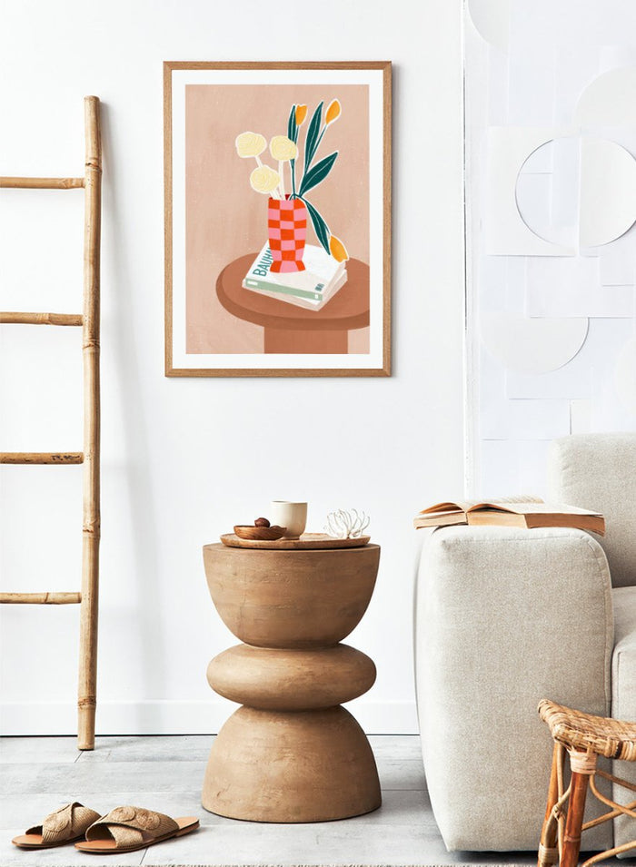 A framed minimalist art print featuring a stylized illustration of a pink and orange checkered vase filled with yellow and orange tulips, resting on a brown round table next to a white book. Colorful Tabletop Flowers by Bea Müller premium framed fine art print featuring yellow tulips and cream blooms in a red-and-pink checkered vase.