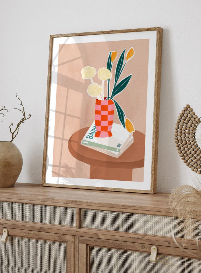 A framed minimalist art print featuring a stylized illustration of a pink and orange checkered vase filled with yellow and orange tulips, resting on a brown round table next to a white book. Colorful Tabletop Flowers by Bea Müller premium framed fine art print featuring yellow tulips and cream blooms in a red-and-pink checkered vase.