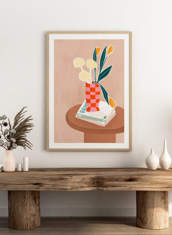 A framed minimalist art print featuring a stylized illustration of a pink and orange checkered vase filled with yellow and orange tulips, resting on a brown round table next to a white book. Colorful Tabletop Flowers by Bea Müller premium framed fine art print featuring yellow tulips and cream blooms in a red-and-pink checkered vase.