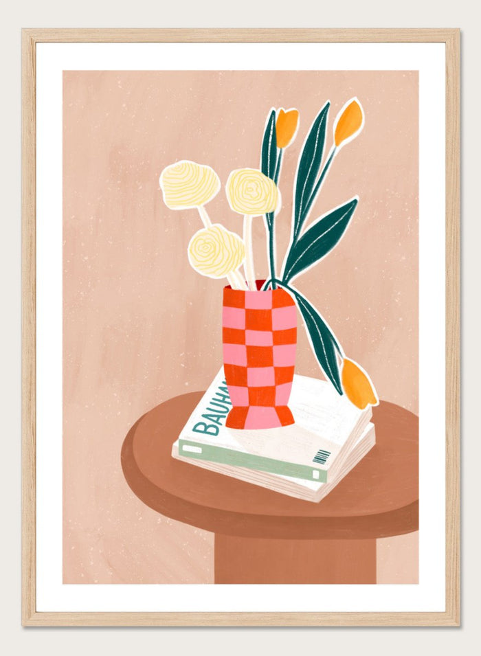 A framed minimalist art print featuring a stylized illustration of a pink and orange checkered vase filled with yellow and orange tulips, resting on a brown round table next to a white book. Colorful Tabletop Flowers by Bea Müller premium framed fine art print featuring yellow tulips and cream blooms in a red-and-pink checkered vase.