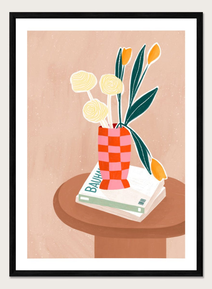 A framed minimalist art print featuring a stylized illustration of a pink and orange checkered vase filled with yellow and orange tulips, resting on a brown round table next to a white book. Colorful Tabletop Flowers by Bea Müller premium framed fine art print featuring yellow tulips and cream blooms in a red-and-pink checkered vase.