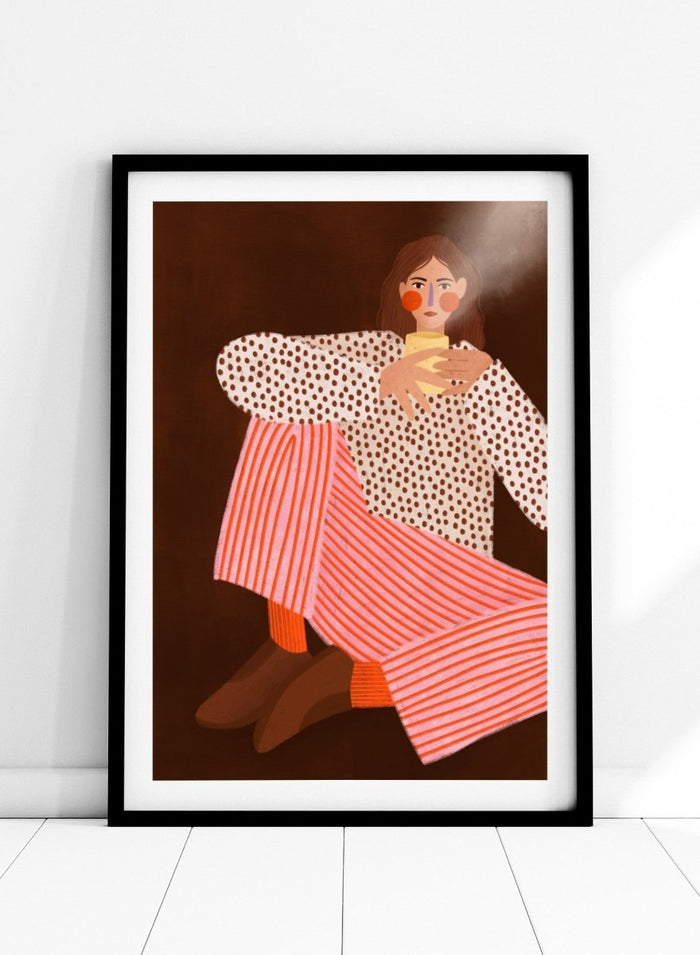 A framed art print featuring a stylized illustration of a woman in a white blouse with brown polka dots and pink-and-white striped trousers, holding a yellow cup against a dark brown background. Cozy Coffee Moment by Bea Müller premium framed fine art print featuring a woman curled up with a warm mug in bold pink, red, and brown tones.