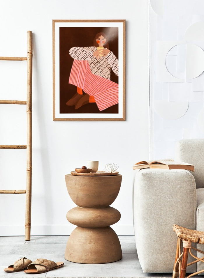 A framed art print featuring a stylized illustration of a woman in a white blouse with brown polka dots and pink-and-white striped trousers, holding a yellow cup against a dark brown background. Cozy Coffee Moment by Bea Müller premium framed fine art print featuring a woman curled up with a warm mug in bold pink, red, and brown tones.