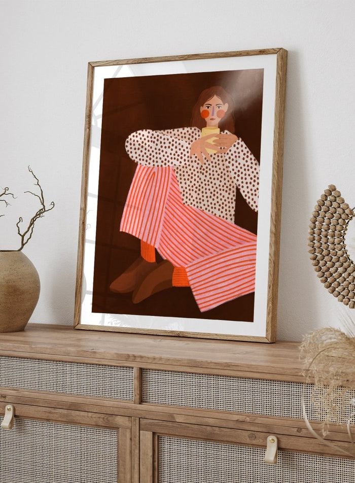 A framed art print featuring a stylized illustration of a woman in a white blouse with brown polka dots and pink-and-white striped trousers, holding a yellow cup against a dark brown background. Cozy Coffee Moment by Bea Müller premium framed fine art print featuring a woman curled up with a warm mug in bold pink, red, and brown tones.