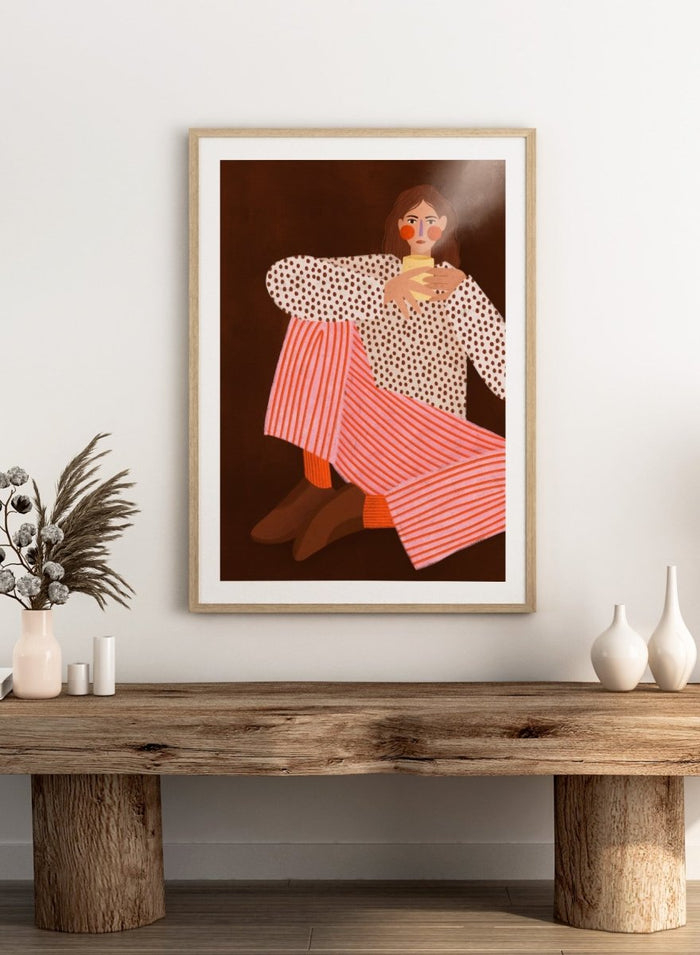 A framed art print featuring a stylized illustration of a woman in a white blouse with brown polka dots and pink-and-white striped trousers, holding a yellow cup against a dark brown background. Cozy Coffee Moment by Bea Müller premium framed fine art print featuring a woman curled up with a warm mug in bold pink, red, and brown tones.