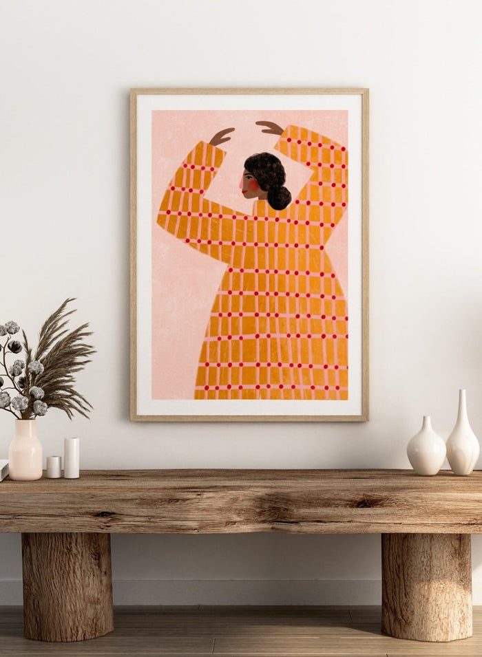A framed art print featuring a stylized illustration of a woman in profile wearing a bright orange dress with a red grid pattern, set against a pale pink background. Dancing Woman in Orange by Bea Müller premium fine art print featuring a bold figure, warm orange tones, and a playful graphic composition.