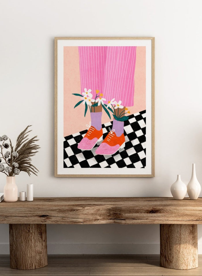 A stylized fashion illustration of a person's legs wearing pink pinstripe trousers, lavender socks, orange and pink shoes, and holding white flowers, set against a peach background with a black-and-white checkered floor. Flowering Oxford Style by Bea Müller premium framed fine art print featuring colorful Oxford shoes, lilac socks, and flowers on a checkered floor.