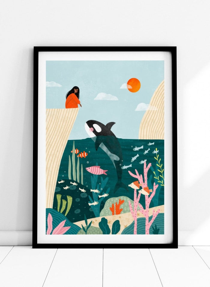 A framed art print featuring a whimsical underwater scene with an orca, coral, fish, and a woman in an orange dress standing on a cliff edge under a sun. Girl Watches Ocean Life by Bea Müller premium framed fine art print featuring a child watching an orca swim through a colorful underwater world.
