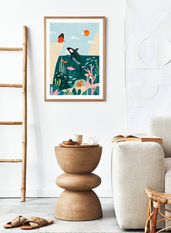 A framed art print featuring a whimsical underwater scene with an orca, coral, fish, and a woman in an orange dress standing on a cliff edge under a sun. Girl Watches Ocean Life by Bea Müller premium framed fine art print featuring a child watching an orca swim through a colorful underwater world.