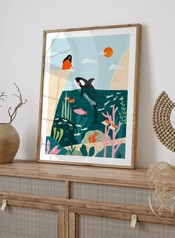 A framed art print featuring a whimsical underwater scene with an orca, coral, fish, and a woman in an orange dress standing on a cliff edge under a sun. Girl Watches Ocean Life by Bea Müller premium framed fine art print featuring a child watching an orca swim through a colorful underwater world.
