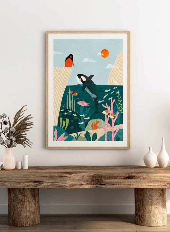 A framed art print featuring a whimsical underwater scene with an orca, coral, fish, and a woman in an orange dress standing on a cliff edge under a sun. Girl Watches Ocean Life by Bea Müller premium framed fine art print featuring a child watching an orca swim through a colorful underwater world.