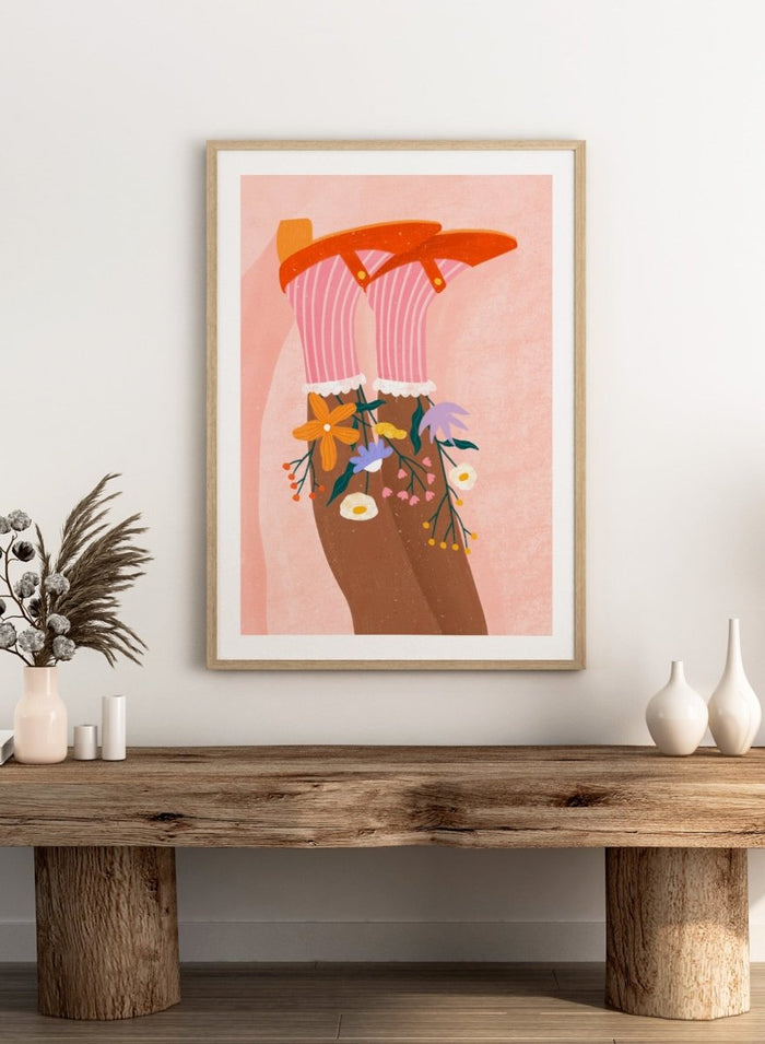 A stylized art print featuring a pair of legs wearing pink striped socks with white ruffles, decorated with colorful wildflowers, and topped with orange high-heeled shoes, set against a textured pink background. Happy Feet and Flowers by Bea Müller premium framed fine art print featuring red Mary Janes, striped socks, and colorful flowers on a soft pink background.