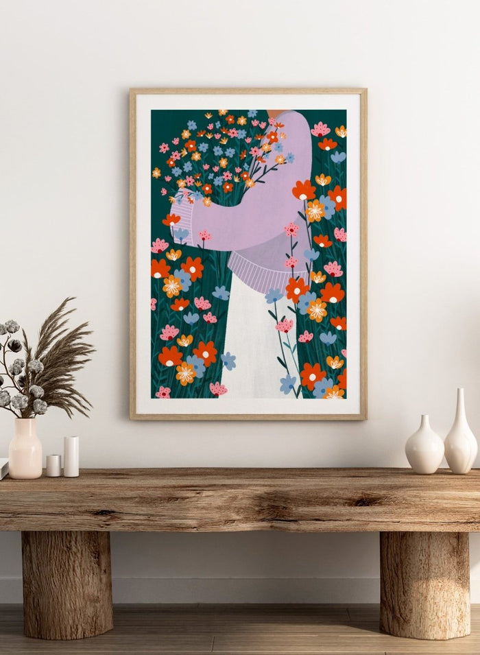 A stylized art print featuring a woman in a lavender sweater and white skirt holding a large bouquet of colorful wildflowers, framed by dense flowers on a dark teal background. Happy Woman Holding Flowers by Bea Müller premium framed fine art print featuring a woman carrying wildflowers in bright orange, pink, blue, and teal.