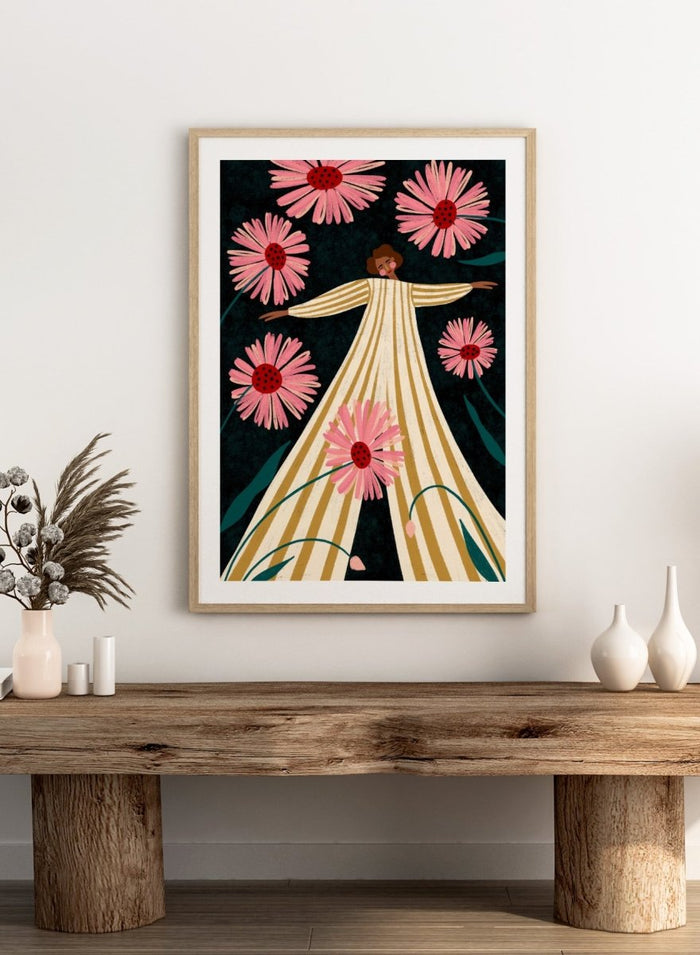 A framed art print featuring a stylized illustration of a woman in a cream and gold striped dress surrounded by large pink flowers on a dark background. Joyful Woman In Bloom by Bea Müller premium framed fine art print featuring a woman in a striped dress surrounded by oversized pink flowers.