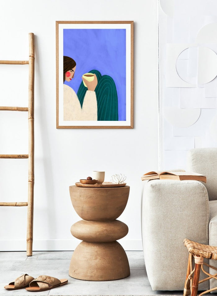 A framed minimalist art print featuring a stylized woman in profile with brown hair, a cream sweater, and a dark teal cape against a textured blue background, holding a pale yellow cup. Peaceful Coffee Moment by Bea Müller premium framed fine art print featuring a woman curled up with a warm drink in soft blue, cream, and teal tones.