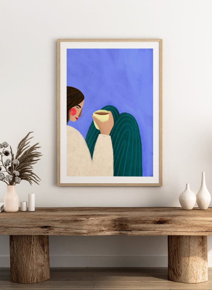 A framed minimalist art print featuring a stylized woman in profile with brown hair, a cream sweater, and a dark teal cape against a textured blue background, holding a pale yellow cup. Peaceful Coffee Moment by Bea Müller premium framed fine art print featuring a woman curled up with a warm drink in soft blue, cream, and teal tones.
