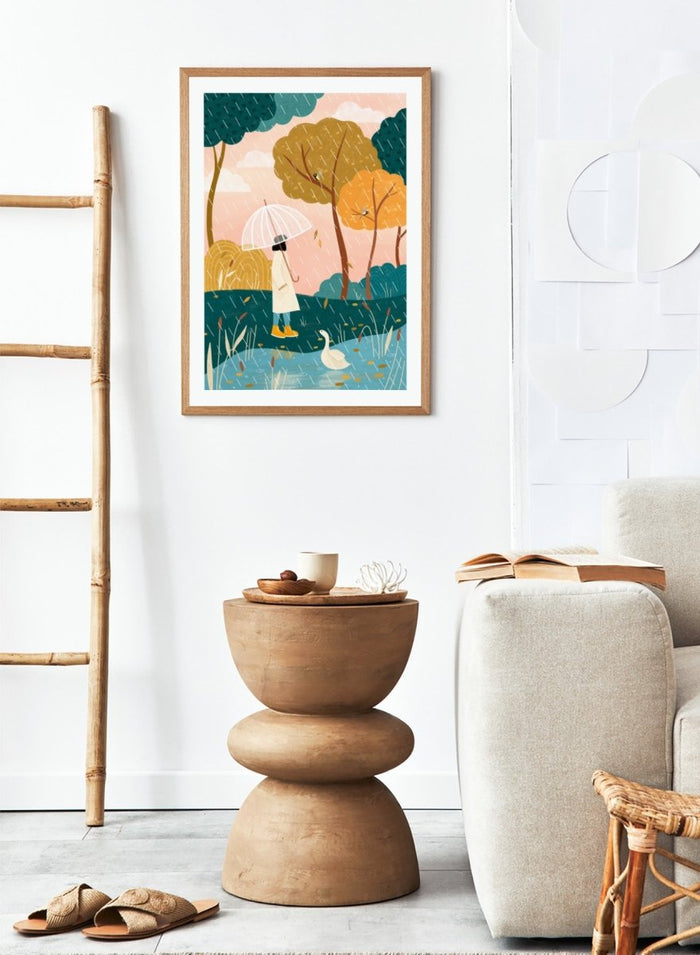 A framed art print featuring a whimsical illustration of a person holding a clear umbrella in the rain near a pond with a white goose, surrounded by stylized trees in teal, mustard, and orange tones. Walking In Autumn Rain by Bea Müller premium framed fine art print featuring a woman with an umbrella beside a pond in a colorful rainy landscape.
