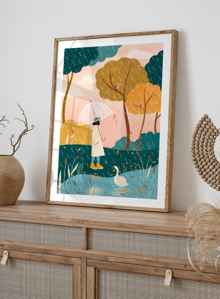 A framed art print featuring a whimsical illustration of a person holding a clear umbrella in the rain near a pond with a white goose, surrounded by stylized trees in teal, mustard, and orange tones. Walking In Autumn Rain by Bea Müller premium framed fine art print featuring a woman with an umbrella beside a pond in a colorful rainy landscape.