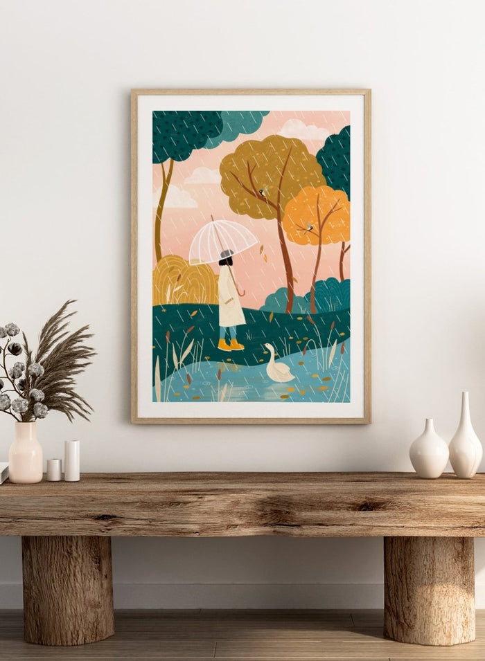 A framed art print featuring a whimsical illustration of a person holding a clear umbrella in the rain near a pond with a white goose, surrounded by stylized trees in teal, mustard, and orange tones. Walking In Autumn Rain by Bea Müller premium framed fine art print featuring a woman with an umbrella beside a pond in a colorful rainy landscape.