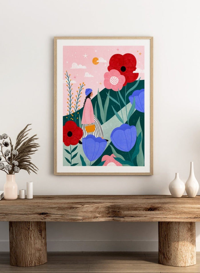 A framed art print featuring a whimsical, colorful illustration of a woman walking through a field of oversized flowers under a pink sky with moon. Whimsical Flower Path by Bea Müller premium fine art print featuring a woman walking among oversized flowers in a bright, playful landscape.