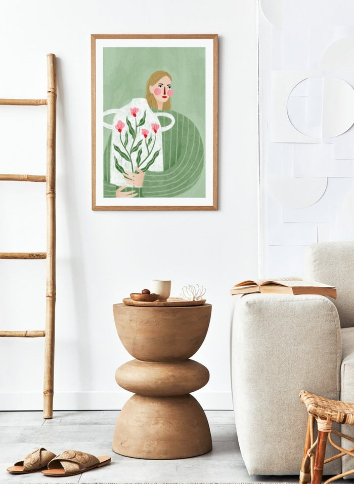 A framed art print featuring a stylized illustration of a woman with blonde hair holding a white apron decorated with pink flowers and green leaves, set against a sage green background. Woman Holding Flowers by Bea Müller premium framed fine art print featuring a woman carrying a white vase filled with pink flowers and green leaves.