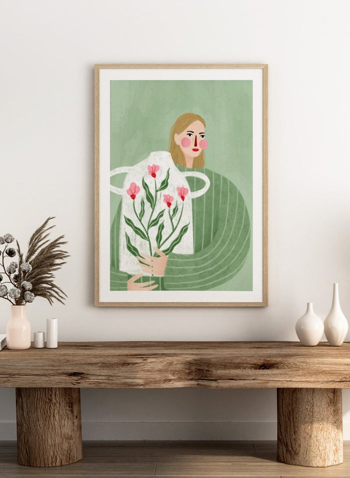A framed art print featuring a stylized illustration of a woman with blonde hair holding a white apron decorated with pink flowers and green leaves, set against a sage green background. Woman Holding Flowers by Bea Müller premium framed fine art print featuring a woman carrying a white vase filled with pink flowers and green leaves.