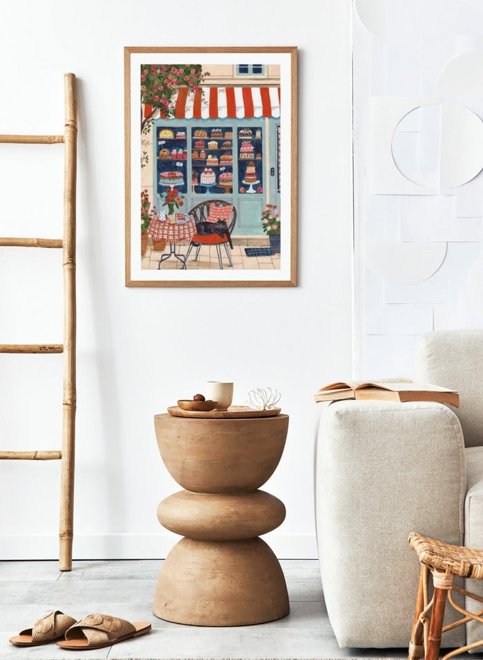 Illustration of a bakery with a cat sitting on a chair outside. Bakery Cat Cafe by Caroline Bonne Müller is a colorful bakery art print featuring cat illustration, pastry shop window, floral details, and modern aesthetic cafe scene.