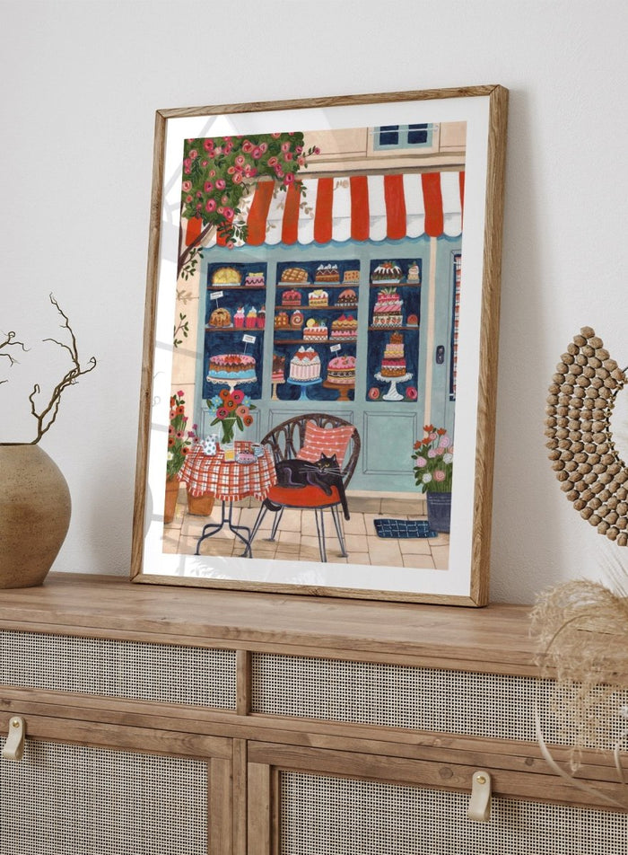 Illustration of a bakery with a cat sitting on a chair outside. Bakery Cat Cafe by Caroline Bonne Müller is a colorful bakery art print featuring cat illustration, pastry shop window, floral details, and modern aesthetic cafe scene.