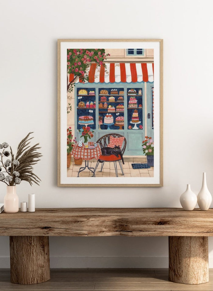 Illustration of a bakery with a cat sitting on a chair outside. Bakery Cat Cafe by Caroline Bonne Müller is a colorful bakery art print featuring cat illustration, pastry shop window, floral details, and modern aesthetic cafe scene.